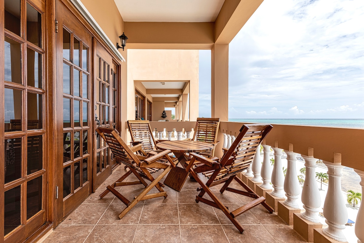 An airy balcony features wooden outdoor furniture, including a round table surrounded by four folding chairs. The view opens to the Caribbean Sea, with the horizon meeting a partially cloudy sky, while decorative white railings enhance the space's elegance.