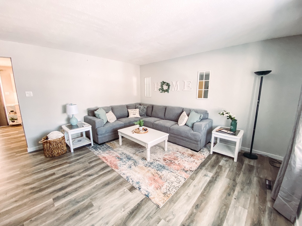 A cozy living room is presented with a large, gray sectional sofa arranged around a light coffee table. Soft lighting from a nearby floor lamp complements the natural light filtering through a window. Decorative accents include a patterned area rug and a small side table with a vase of flowers.