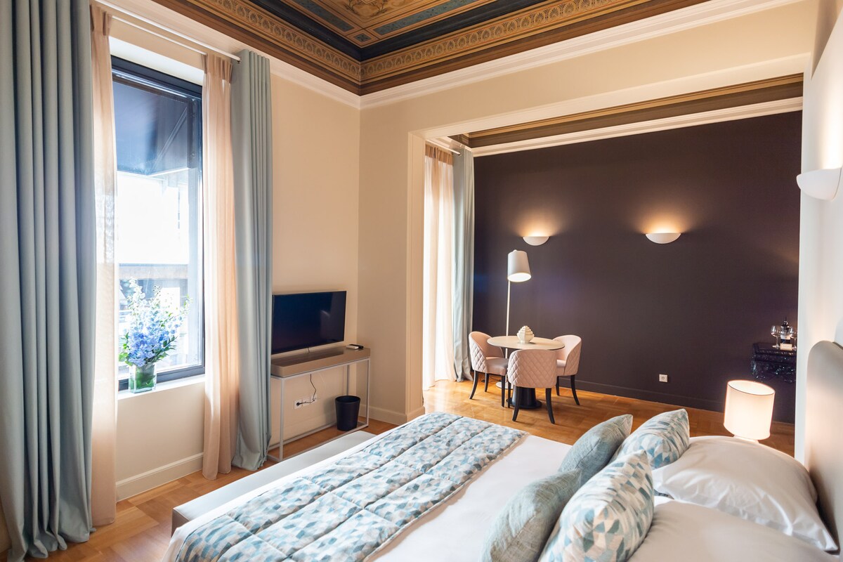 A spacious bedroom features high hand-painted ceilings and wooden floors. A cozy queen-sized bed with a patterned duvet is complemented by a seating area with a round table and two chairs. Natural light floods in through a large window, with soft curtains framing the view.