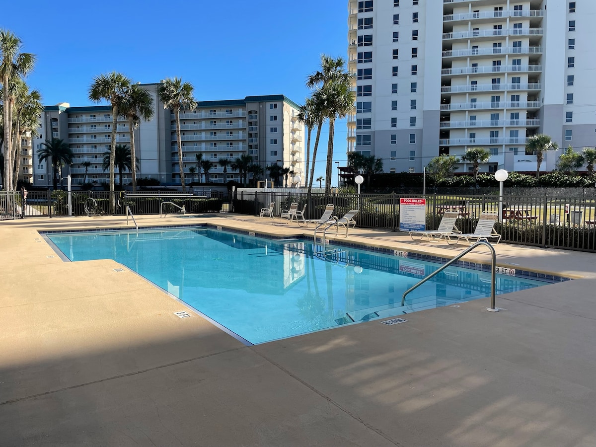 A sparkling outdoor pool is framed by palm trees and sun loungers, with a clear blue sky above. Surrounding buildings include two multi-story condos, adding a sense of community. The pool area is well-maintained, providing a relaxing retreat for guests.