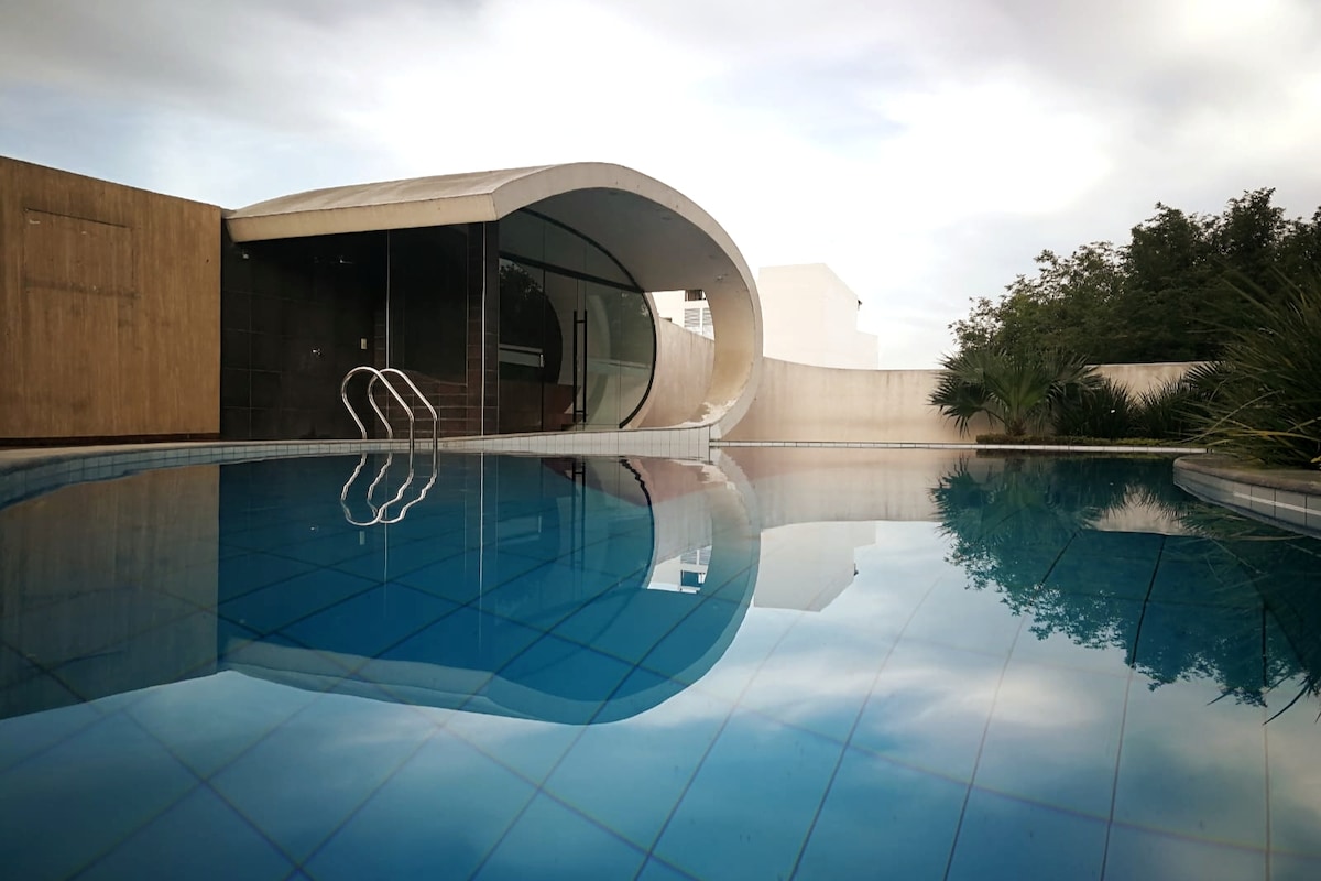 A tranquil swimming pool reflects the modern architecture nearby. The pool features clear blue water, with steps leading into it. Surrounding greenery and a gentle curve of the building complement the serene outdoor space.