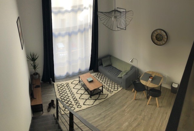 A spacious living area is highlighted by large windows allowing natural light to fill the room. A comfortable sofa and a coffee table are positioned on a patterned rug, while a small dining table with chairs is situated nearby. Decorative elements include a hanging light fixture and wall art.