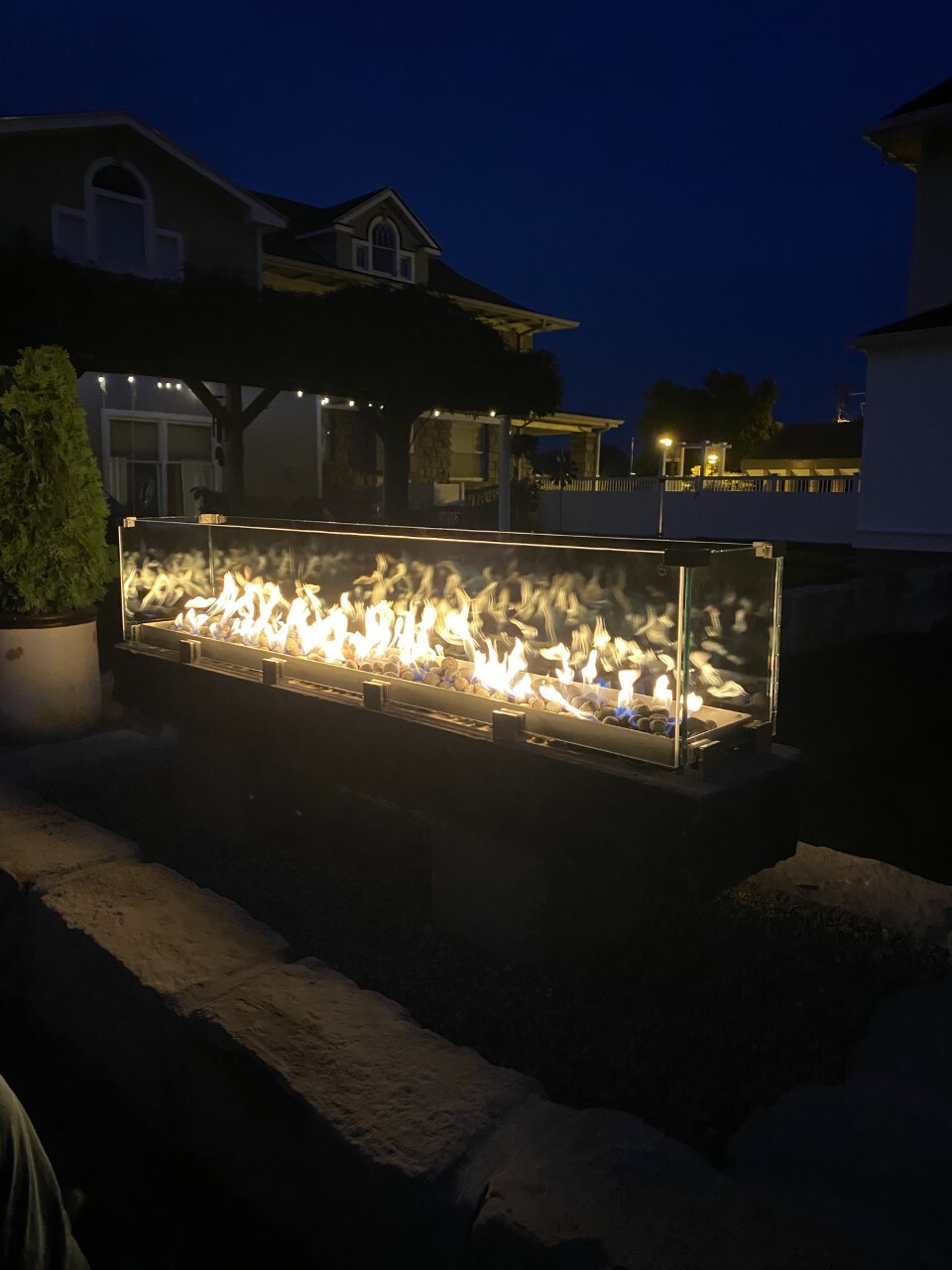 An outdoor fire feature is prominently displayed, illuminated against the evening sky. The flames dance behind a glass barrier, creating a warm glow. Lush greenery and structures can be seen in the background, adding to the relaxing ambiance of the outdoor space.