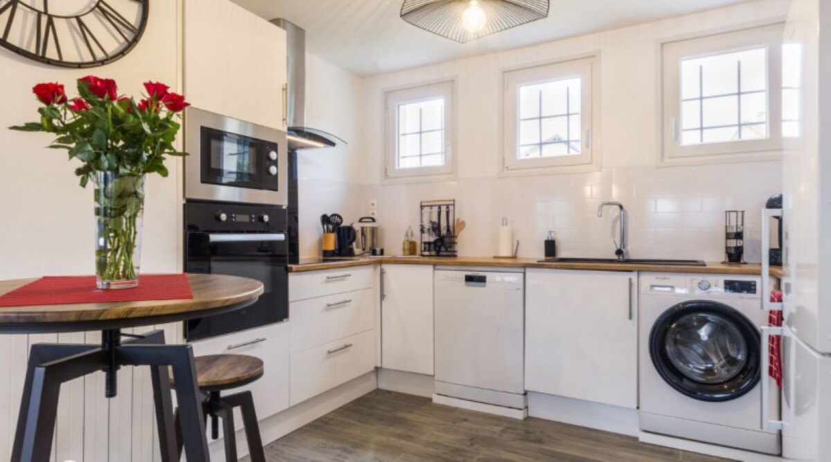 The modern kitchen is equipped with essential appliances, including a dishwasher and washing machine. A small table with two stools is positioned beside a vase of roses. Natural light enters through three square windows, illuminating the space.