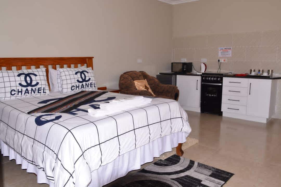 A spacious sleeping area is highlighted by a large bed adorned with a grid-patterned blanket featuring logo designs. A cozy armchair is positioned near a kitchenette, which includes a microwave and stove, providing essential amenities for guests' convenience.