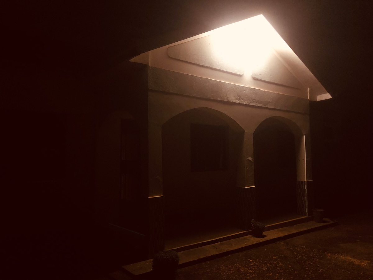 An illuminated house stands under the night sky, highlighting its arched entryway and subtle architectural features. Soft light emanates from above the porch, casting gentle shadows on the surroundings. The area appears quiet and serene, inviting a peaceful atmosphere.