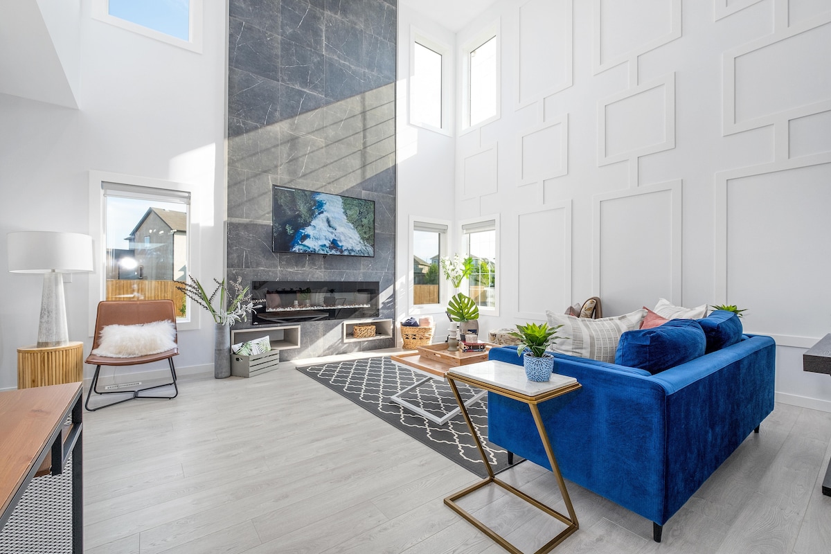 A spacious living room features high ceilings, large windows allowing natural light, and a modern fireplace. A blue sofa faces a sleek wall-mounted TV, complemented by a geometric rug and potted plants. Additional seating is provided by a stylish chair, enhancing the inviting setting.