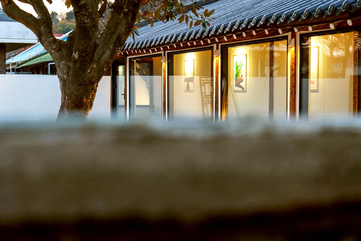 The image captures a view of a traditional Hanok structure, showcasing lit windows framed by wooden beams. A tree stands in the foreground, adding a natural element. The peaceful ambiance is enhanced by the surrounding architecture, making the area feel serene and inviting.