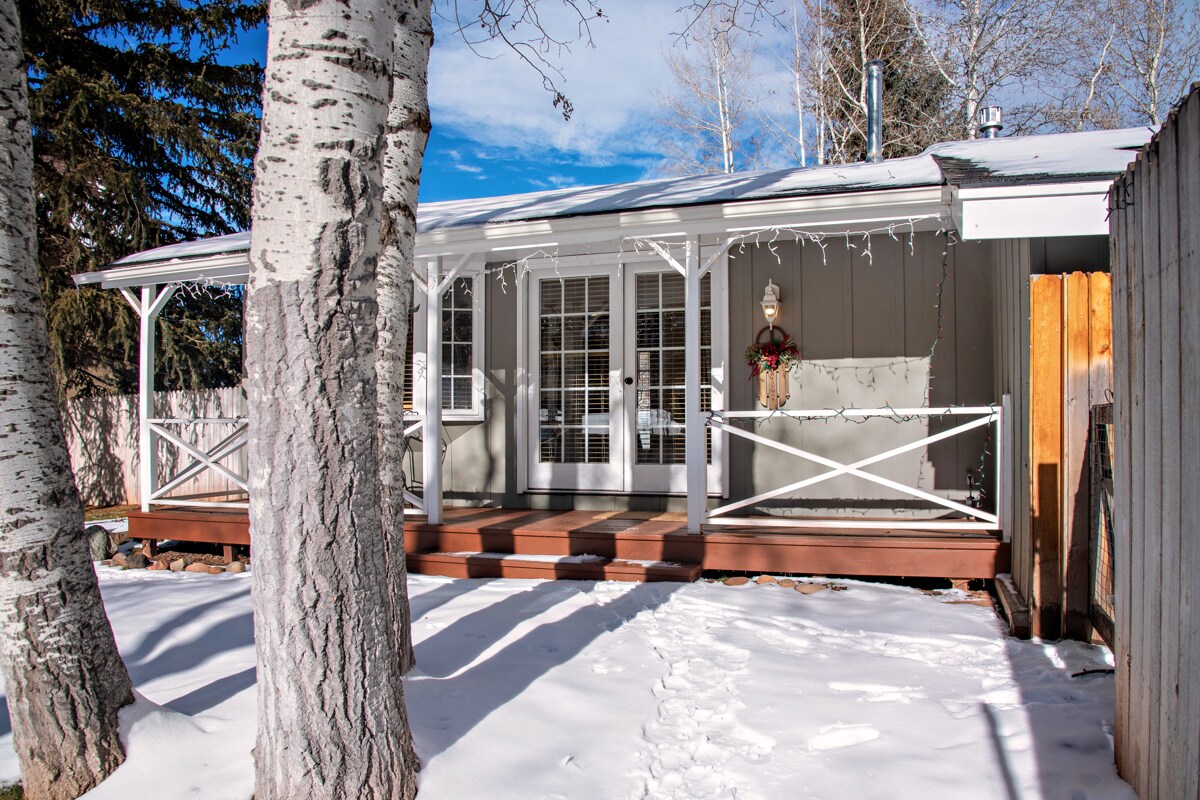 The front porch of a cozy cottage features wooden decking and a railing, warmly illuminated by string lights. Snow blankets the ground, while tall, white bark trees frame the entrance. French doors open to the interior, enhancing the inviting atmosphere.