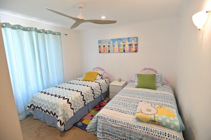 تحتوي غرفة النوم المريحة هذه على سريرين مفردين مع مفارش مبهجة ومنقوشة ووسائد ملونة. توجد طاولة صغيرة بيضاء بين السريرين، وتضيف الستائر المائية الناعمة لمسة مهدئة. يعزز فن الحائط المرح بتصميم لوح التزلج على الماء من الاسترخاء