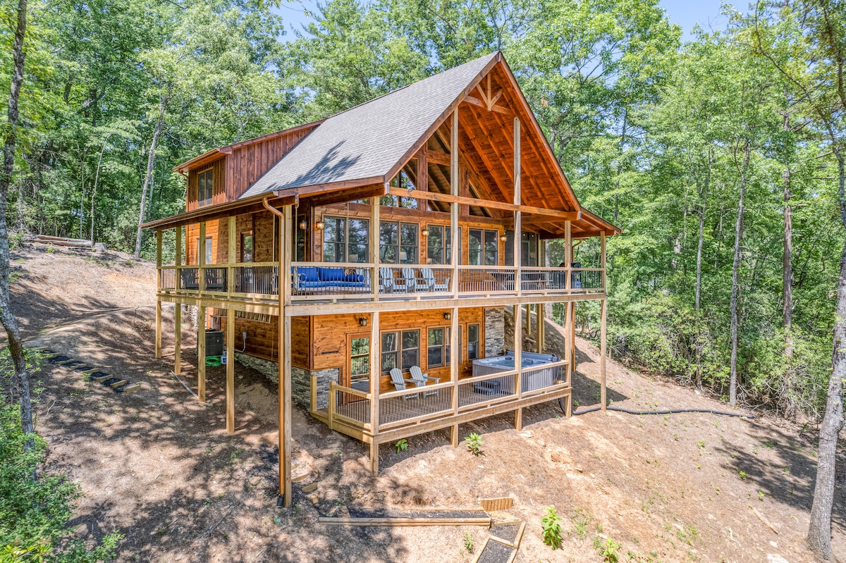 The cabin features a bright and spacious exterior with multiple levels. Large windows invite natural light, while expansive decks provide areas for seating and relaxation. Surrounded by trees, the setting highlights the cabin's connection to nature.
