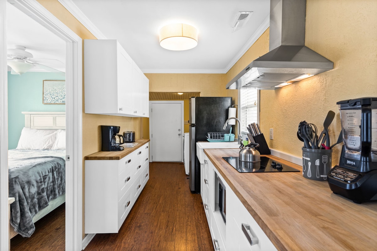 A modern kitchen space is shown, featuring white cabinetry and wooden countertops. A steel hood is positioned above a stovetop. Small appliances like a coffee maker and blender are visible. Natural light enters through a window, illuminating the bright yellow walls.