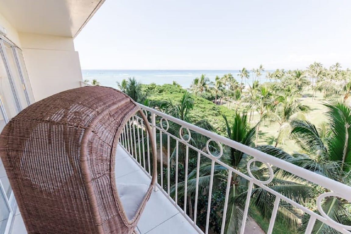 A lanai with a chic rattan chair is presented, overlooking lush greenery and the ocean in the distance. The railing is white, complementing the serene natural setting and enhancing the view of palm trees swaying gently.
