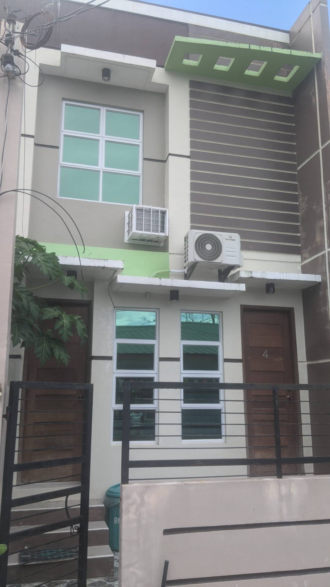 The exterior of the building is characterized by a modern facade with neutral tones and vibrant green accents. Two doors are visible, each with distinct wooden finishes. A window above each door allows natural light in, while an air conditioning unit is mounted on the wall.