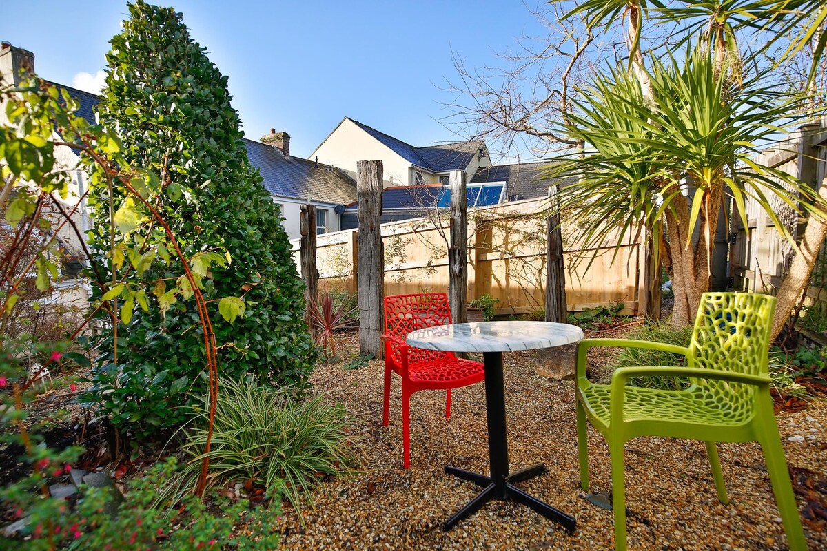 A private garden area features a round table with four legs, surrounded by two brightly colored chairs in red and green. Lush greenery forms a backdrop, including a tall shrub and palm-like plants, set against the warm tones of the cottage.