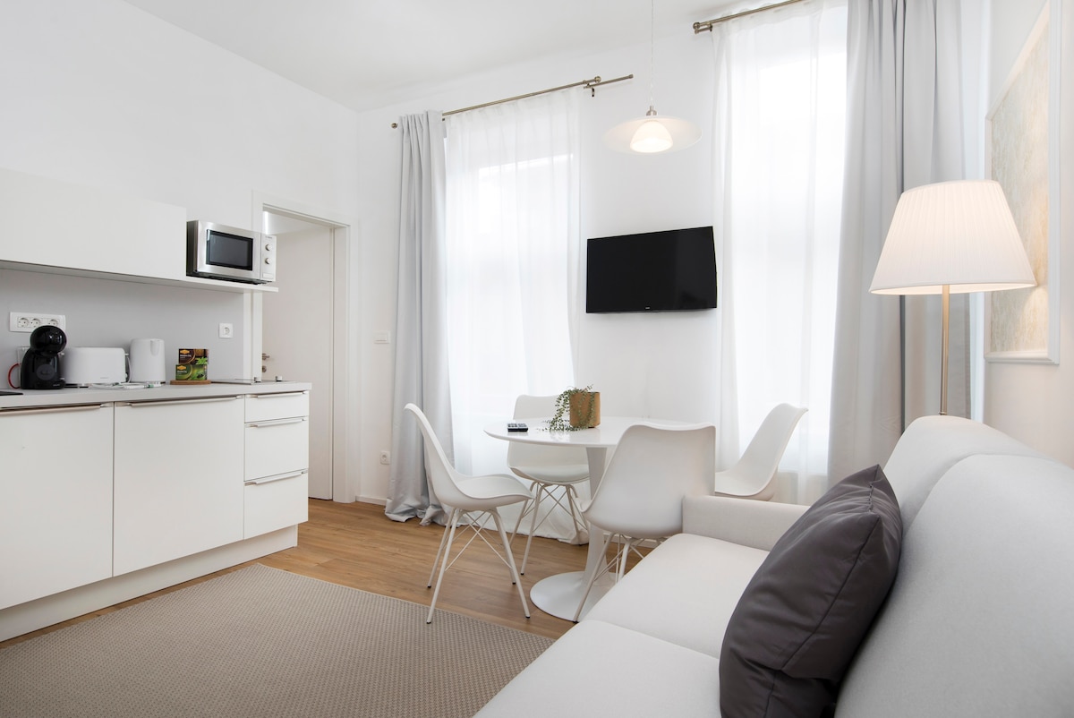 The living area showcases a spacious layout with a light-colored sofa and a sleek white dining table surrounded by modern chairs. Natural light enters through tall windows, complemented by soft curtains. A wall-mounted television and a minimalist light fixture enhance the contemporary design.