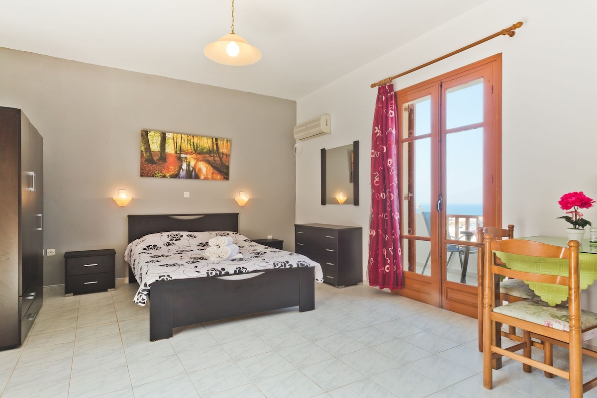 A spacious room features a large bed covered with a patterned blanket and flanked by bedside lamps. Natural light enters through double doors leading to a balcony, while a dining table and chairs provide a comfortable seating area. A scenic landscape painting adds a touch of decor.