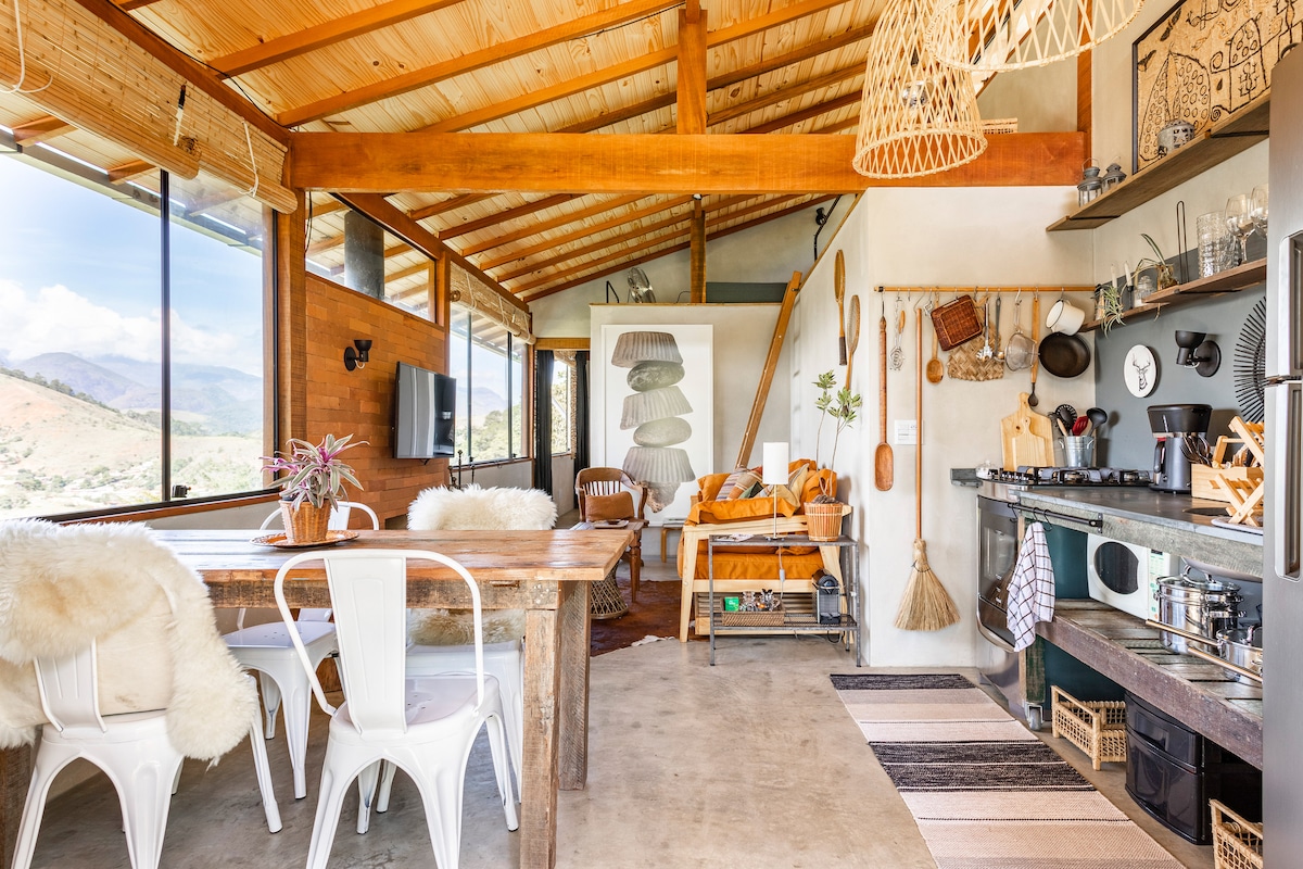 An open living space features wooden beams and large windows that frame mountain views. A rustic dining table is surrounded by white chairs, while a kitchen is equipped with essential appliances and quality utensils. Soft textiles and natural elements provide a cozy ambiance.