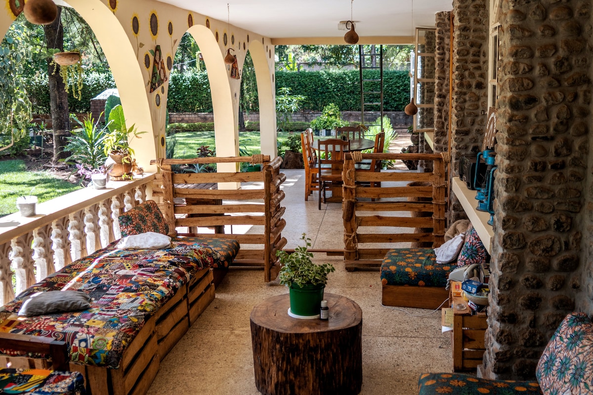 An outdoor seating area is surrounded by archways, featuring colorful cushions on wooden benches and a central round table. Lush greenery can be seen in the background, enhancing the inviting environment. Natural light brightens this communal space, ideal for relaxation and conversation.