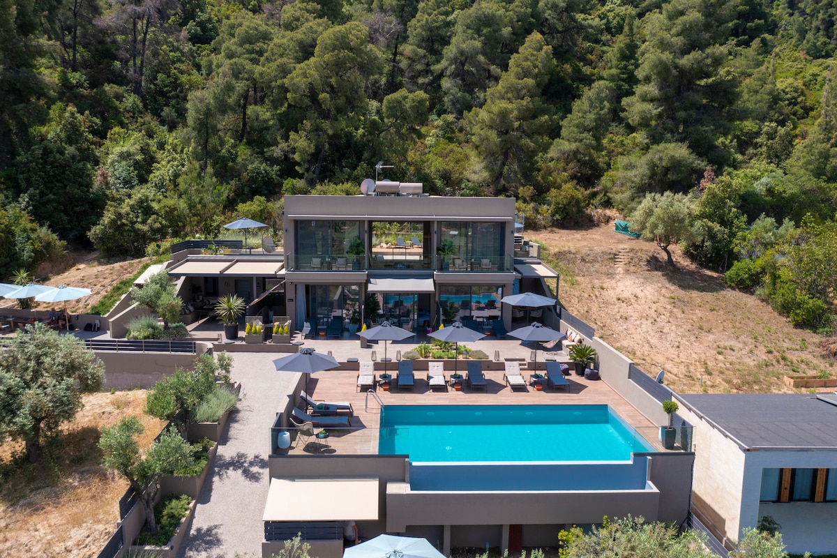 The property features a modern, multi-level structure surrounded by lush greenery. An inviting infinity pool sits at the forefront, accompanied by lounging areas with sun umbrellas. Large glass doors showcase views of the interior and outdoor spaces, blending seamlessly with the natural landscape.