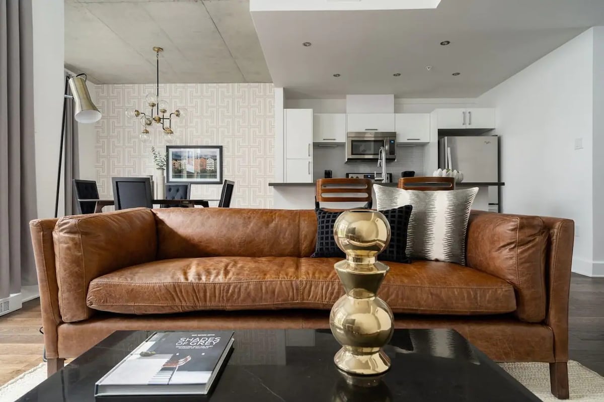A stylish living area features a brown leather sofa paired with decorative cushions. A coffee table sits in front, displaying a book. The background includes a dining area with a dark wooden table and chairs, while the kitchen showcases modern appliances.