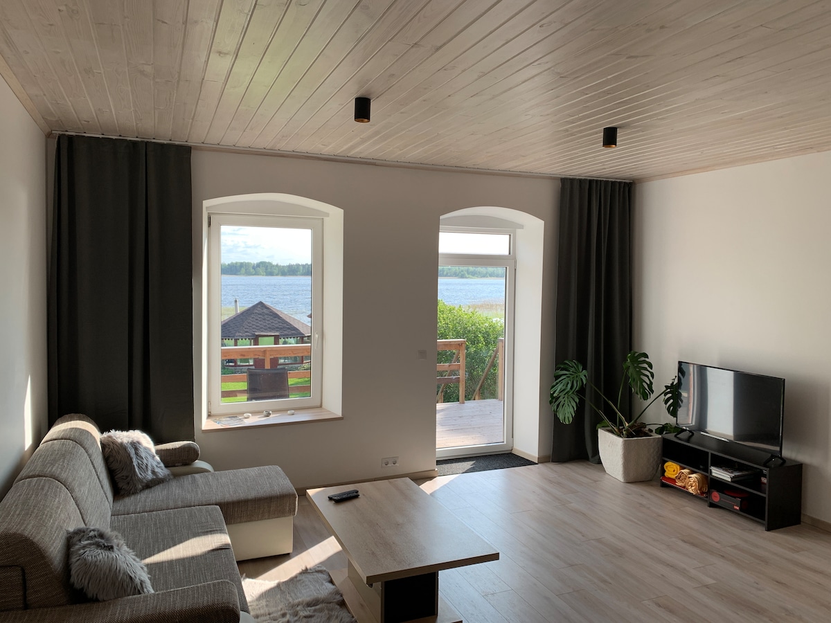 A spacious living area features large windows offering views of the nearby lake. A comfortable sofa is arranged around a central coffee table, with a TV positioned on a console against the wall. Light wood paneling and neutral tones create a calming environment.