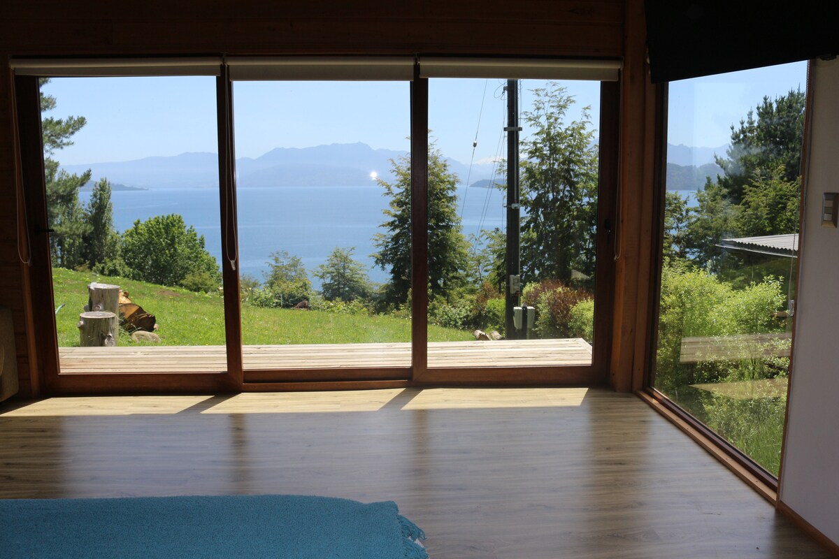 Large glass doors provide an unobstructed view of the lake and mountains, inviting natural light into the space. A smooth wooden floor extends towards the outdoor area, which is surrounded by greenery. The serene landscape is enhanced by the presence of lush trees.