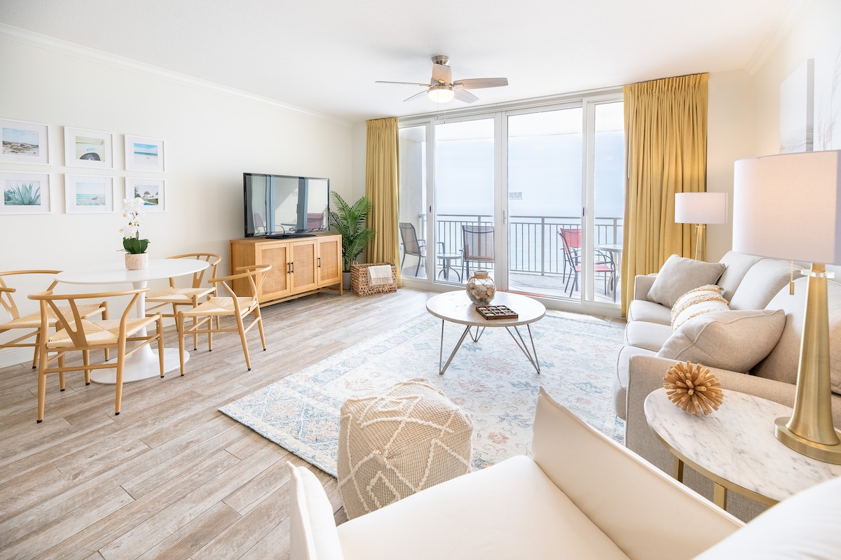 A bright and open living area features large glass doors leading to a spacious balcony with ocean views. Soft furnishings include a light-colored sofa and armchair, paired with a round coffee table. A dining table for four and stylish decor elements complete the inviting space.