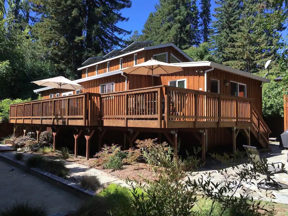 A wooden deck extends from the house, providing multiple shaded areas with umbrellas. The home is surrounded by lush greenery and tall trees, creating a shaded outdoor environment. Stone pathways and landscaped gardens enhance the tranquil, natural setting.