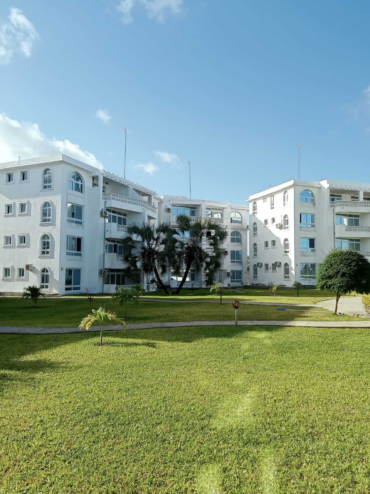 The exterior of a white multi-story apartment building is framed by manicured lawns and palm trees. Large arched windows reflect the blue sky, and balconies are visible on multiple levels. The environment appears calm and well-maintained, contributing to a welcoming atmosphere.