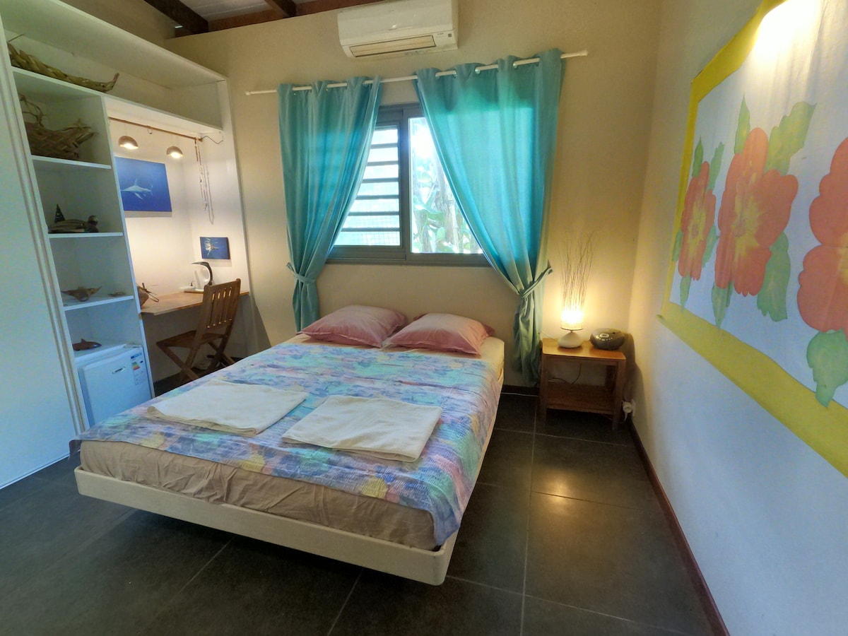 A cozy bedroom features a spacious bed adorned with colorful linens. Light enters through a window covered by sheer blue curtains, illuminating the room. A small desk is positioned nearby, complemented by a shelf displaying decorative items.