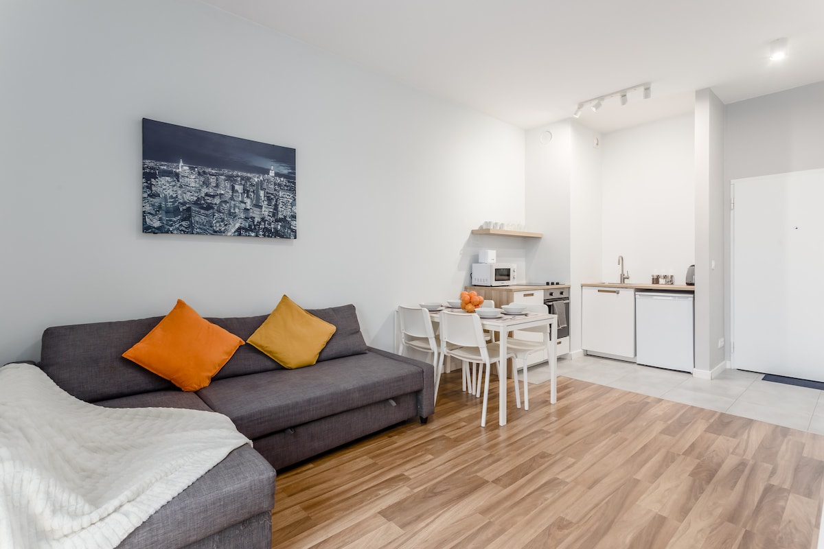A modern living space features a gray sofa with orange and yellow pillows, complemented by a dining area with white chairs and a table. A small kitchen area with appliances is visible in the background, and a cityscape artwork adorns the wall.