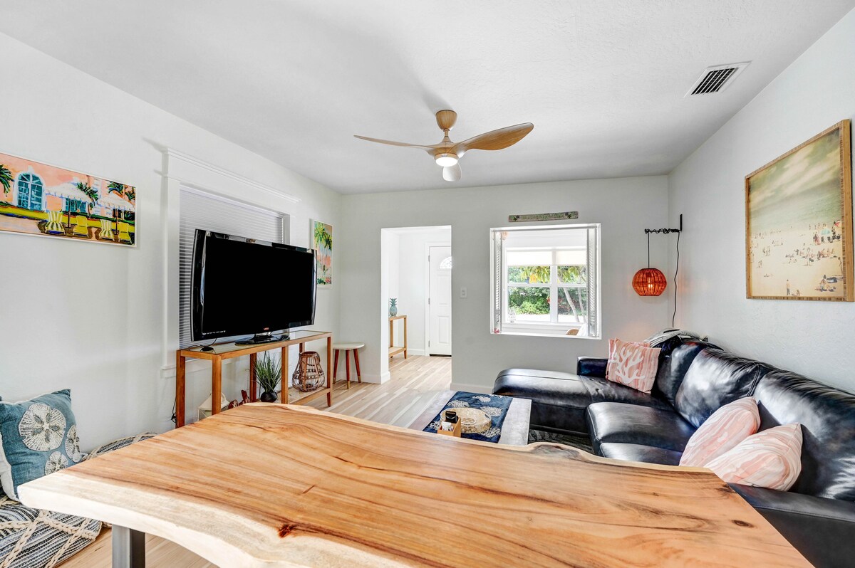 A bright living area features a large wooden table, a sleek black sofa, and a wall-mounted TV. Colorful artwork adorns the walls, and a ceiling fan provides airflow. Natural light streams through a window, illuminating the space.