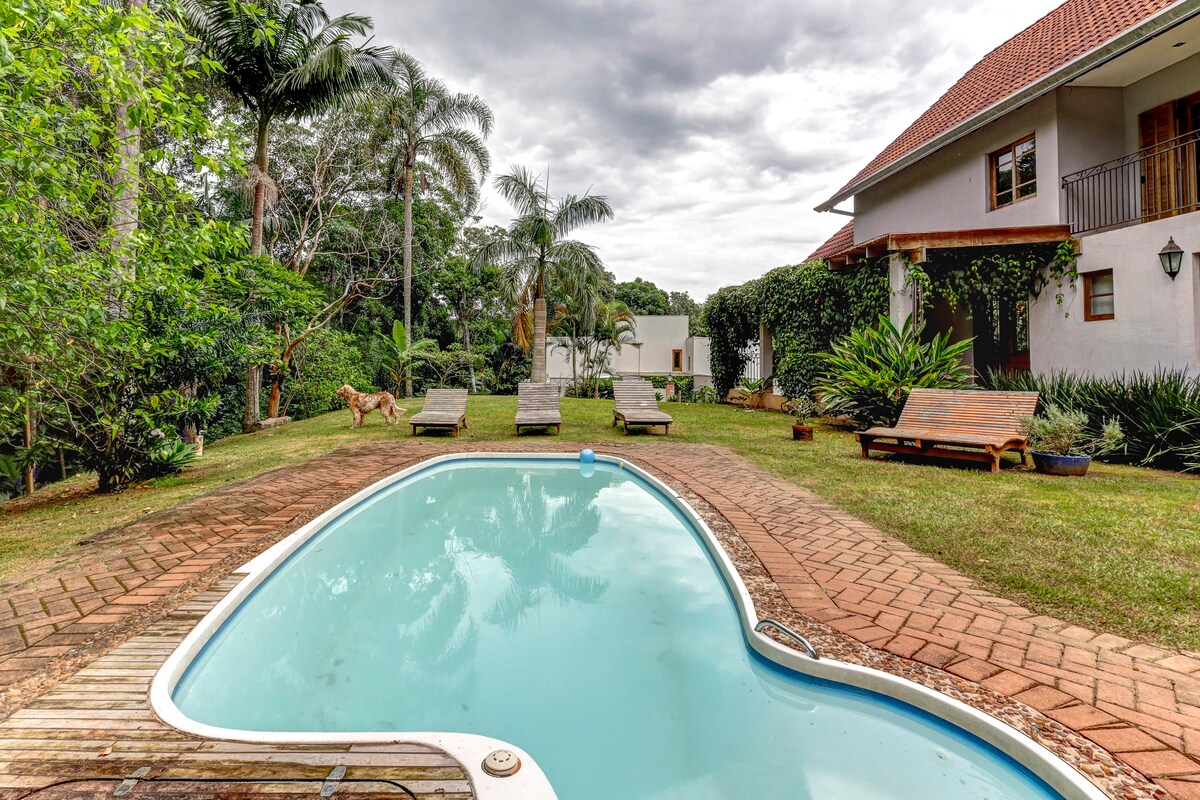 A tranquil pool is surrounded by a well-maintained lawn, with lounge chairs positioned for relaxation. Lush greenery and tropical trees create a serene backdrop, while a spacious white building is visible nearby, highlighting a harmonious blend of nature and comfort.