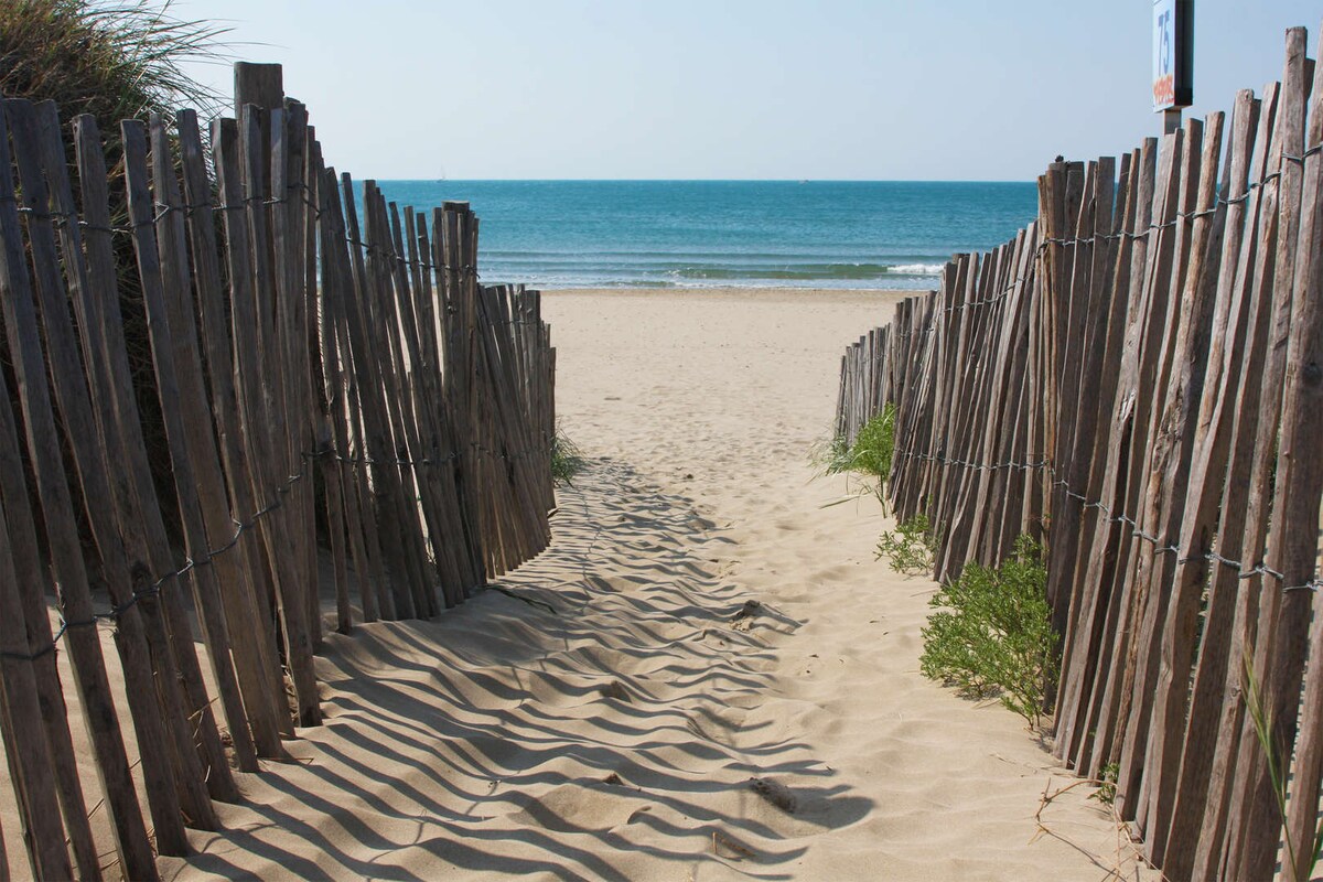 A sandy pathway leads to a serene beach, framed by rustic wooden fencing on both sides. Gentle waves can be seen in the distance under a clear sky. Soft shadows are cast in the sand, enhancing the tranquil scene.