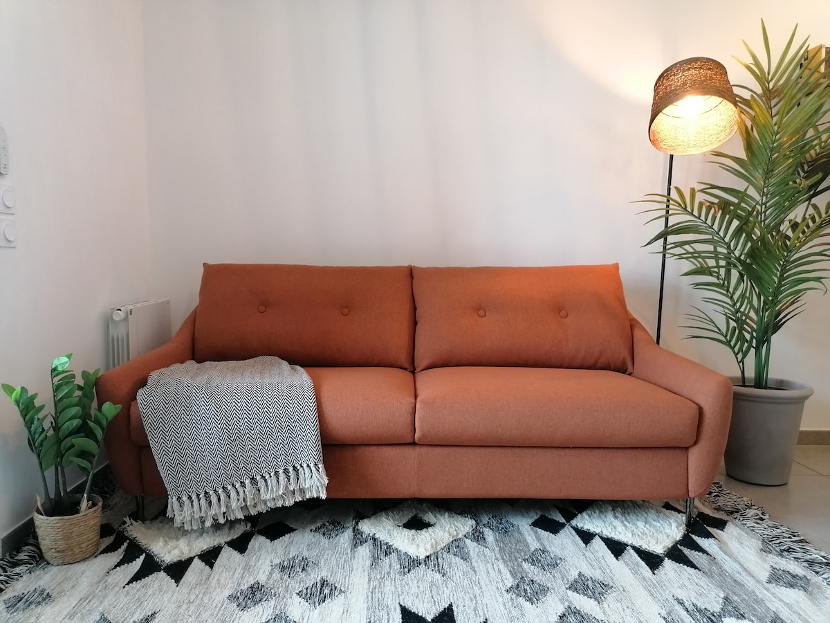 A spacious brown sofa features a clean design with distinct buttons and a soft throw draped over one arm. Nearby, a potted plant adds a touch of greenery, and a decorative lamp provides warm lighting, enhancing the cozy atmosphere of the living area.