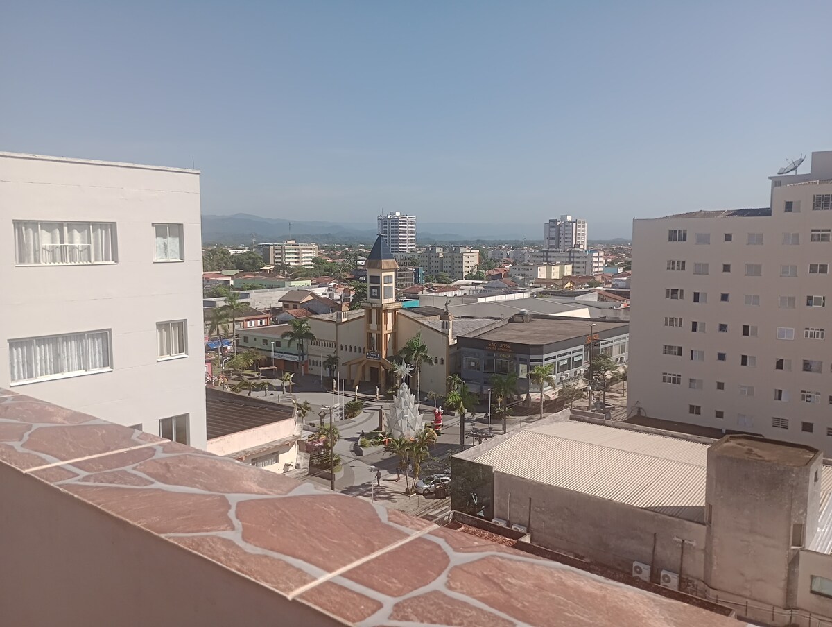 A cityscape view is presented from a balcony, featuring a church with a distinct clock tower in the foreground. Surrounding buildings showcase a mix of residential and commercial structures. The horizon is framed by distant mountains under a clear blue sky.