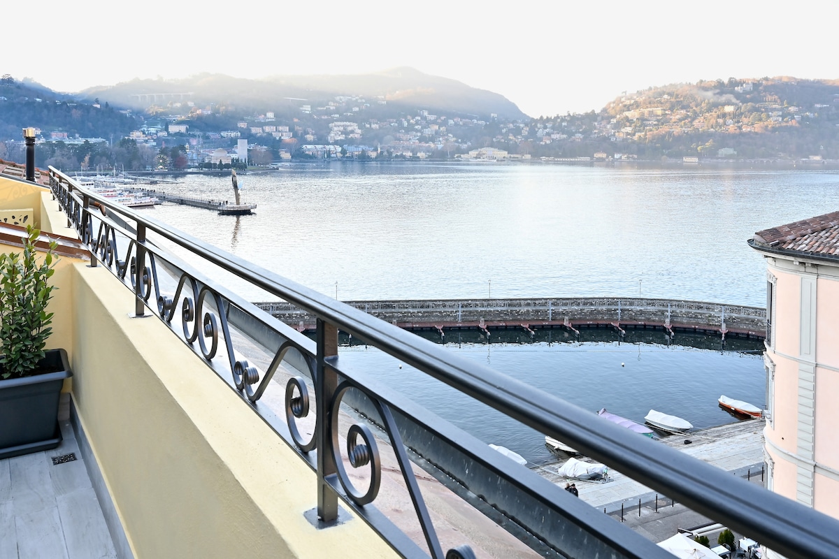 A balcony railing with ornate metalwork overlooks a serene lake, with gentle waves reflecting the surrounding hills and a town in the distance. Small boats are visible along the shore, adding to the tranquil lakeside scenery.