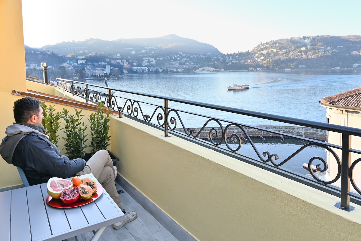 A balcony offers serene views of the lake, framed by decorative railings. A small table is set with fresh fruit, while a person relaxes in a chair, enjoying the peaceful ambiance of the surroundings. Boats can be seen gently gliding across the water.