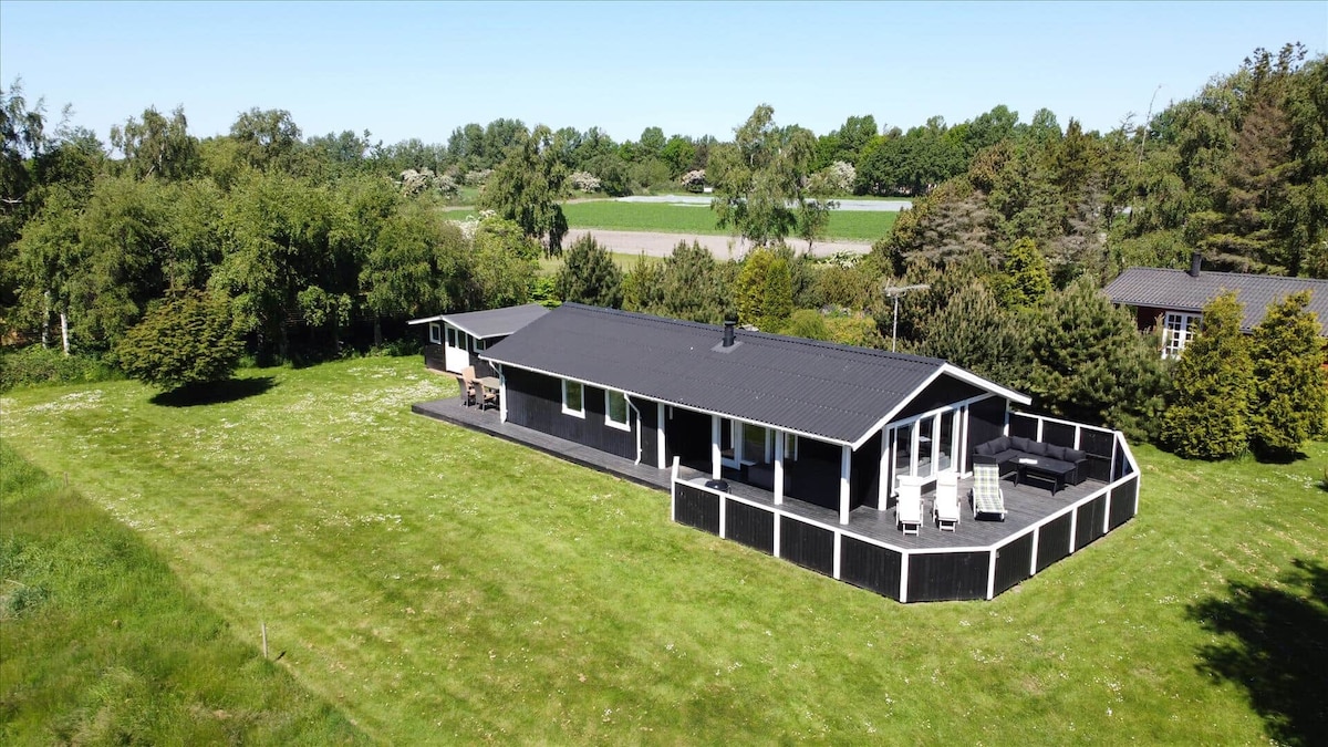 A spacious holiday home is shown nestled within a large grassy area, surrounded by lush greenery. The structure features a dark roof and white siding, with multiple outdoor seating areas visible on the expansive patio. The surrounding landscape is open and serene.