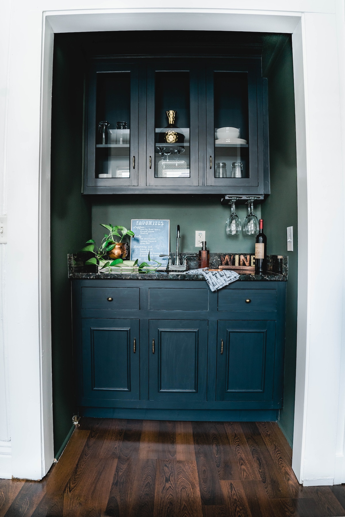 A small kitchenette is set within a recessed alcove, featuring dark cabinetry with glass doors. Glassware and dishes are neatly arranged inside. A countertop is equipped with a wine bottle and accessories, alongside a potted plant for a touch of greenery.