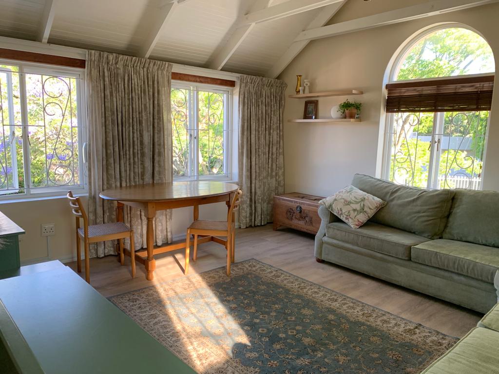 A bright lounge area features large windows framed by soft curtains, allowing natural light to fill the space. A wooden dining table with two chairs stands alongside a comfortable green sofa. A decorative shelf adorns the wall, adding a touch of charm.