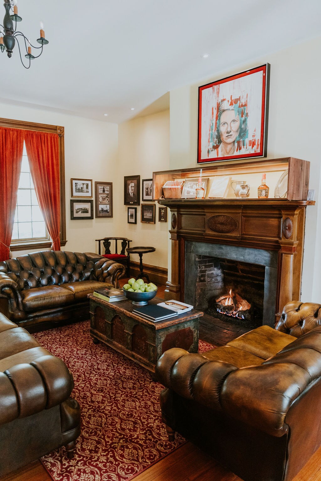 A cozy sitting area features plush leather sofas arranged around a wooden coffee table. A decorative fireplace serves as a focal point, complemented by family photographs on the walls. Rich red curtains frame a window, enhancing the room's warm ambiance.