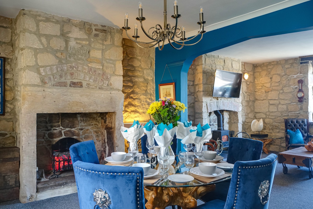 A charming dining area is presented with a round table set for a meal, featuring elegant tableware. A decorative fireplace with a warm glow is visible, complemented by exposed stone walls. A chandelier adds light to the room, enhancing its welcoming ambiance.