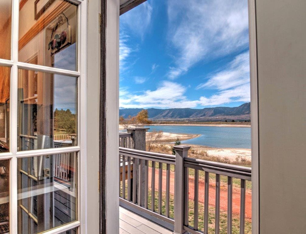 A view from a doorway showcases a sweeping landscape of water and rolling hills, framed by a railing. The sky features clouds and bright blue tones, highlighting the tranquility of the lakeside setting.