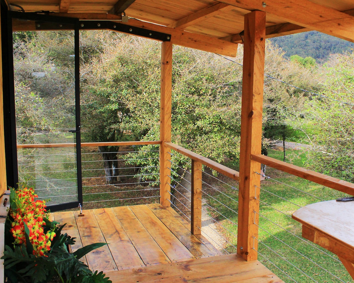 An outdoor balcony area is framed by wooden beams, featuring a wooden floor. A small table is positioned near the railing, while potted plants with vibrant blossoms add color. Lush greenery is visible in the background, providing a serene connection to nature.