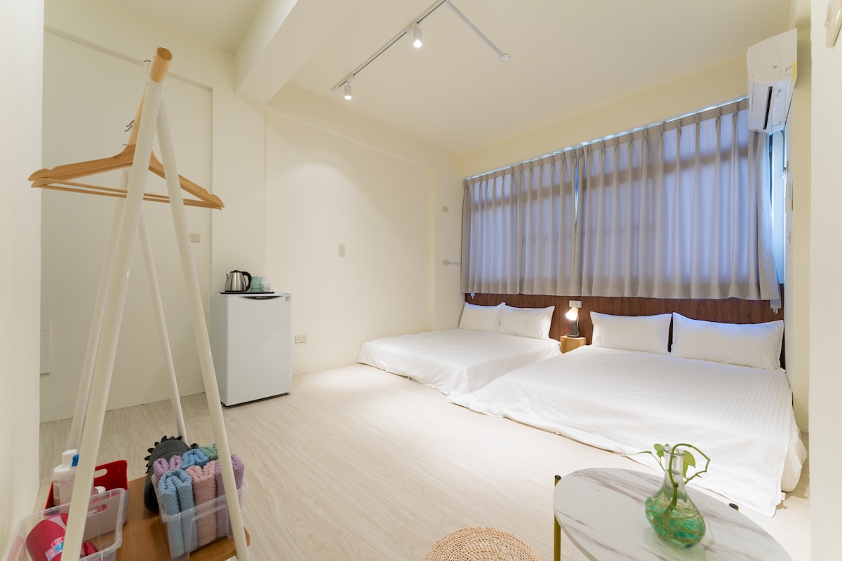 A spacious room features two futon-style beds against a soft-colored wall. Natural light filters through sheer curtains, illuminating the light wood flooring. A small refrigerator and kettle are positioned in the corner, while a minimalist clothing rack adds functionality to the space.