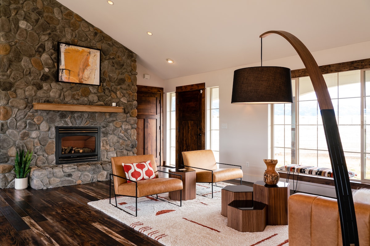 The living room features a stone fireplace, enhancing the modern rustic aesthetic. Two tan lounge chairs with patterned cushions sit adjacent to wooden tables. A large floor lamp provides illumination, while large windows allow natural light and views of the surrounding landscape.