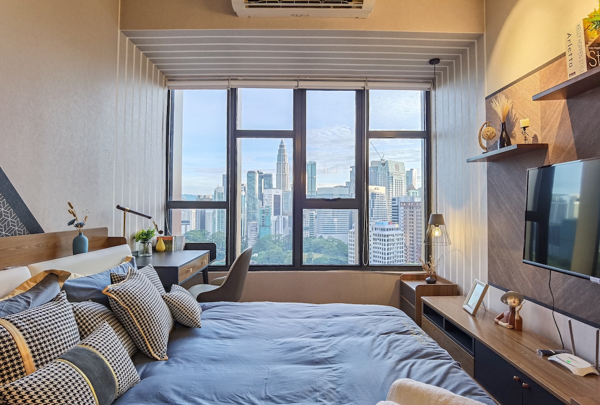 A private studio room features a spacious bed adorned with patterned cushions. Large windows frame a view of the Kuala Lumpur skyline, allowing natural light to illuminate the space. A desk and television are positioned on the opposite wall, enhancing functionality.