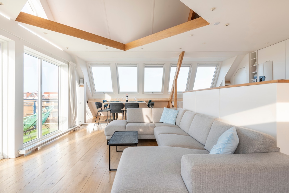 A bright living area is highlighted by a spacious light gray sectional sofa and a sleek black coffee table. Large windows allow natural light to fill the room, while a dining area with a table and chairs is positioned in the background, offering a view of the outside.